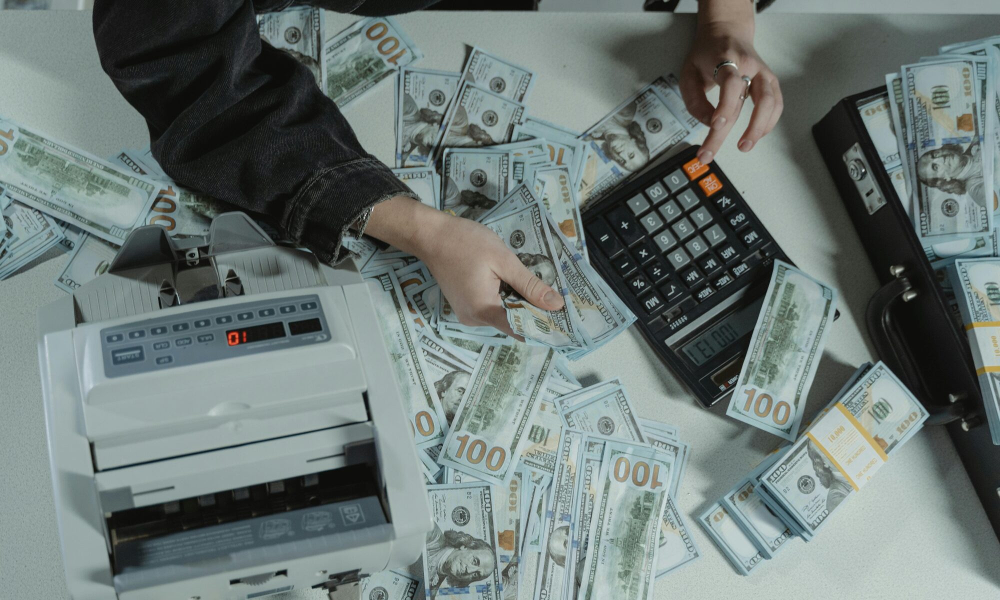Hands counting 100 dollar bills using a calculator and money counter on a table.