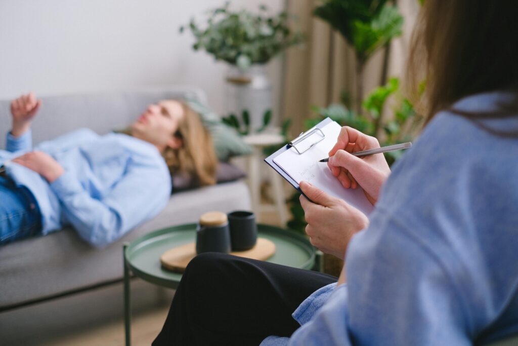 Unrecognizable professional therapist writing on clipboard while sitting against male client lying on couch on blurred background during mental health therapy