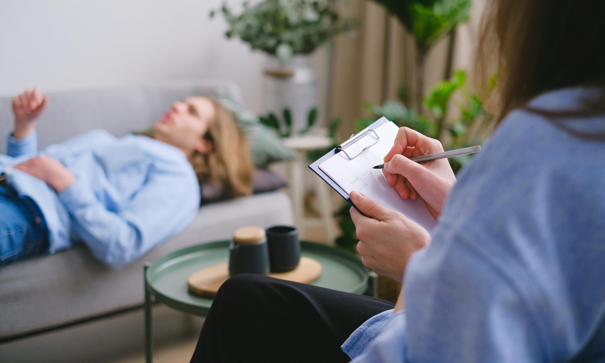 Unrecognizable professional therapist writing on clipboard while sitting against male client lying on couch on blurred background during mental health therapy
